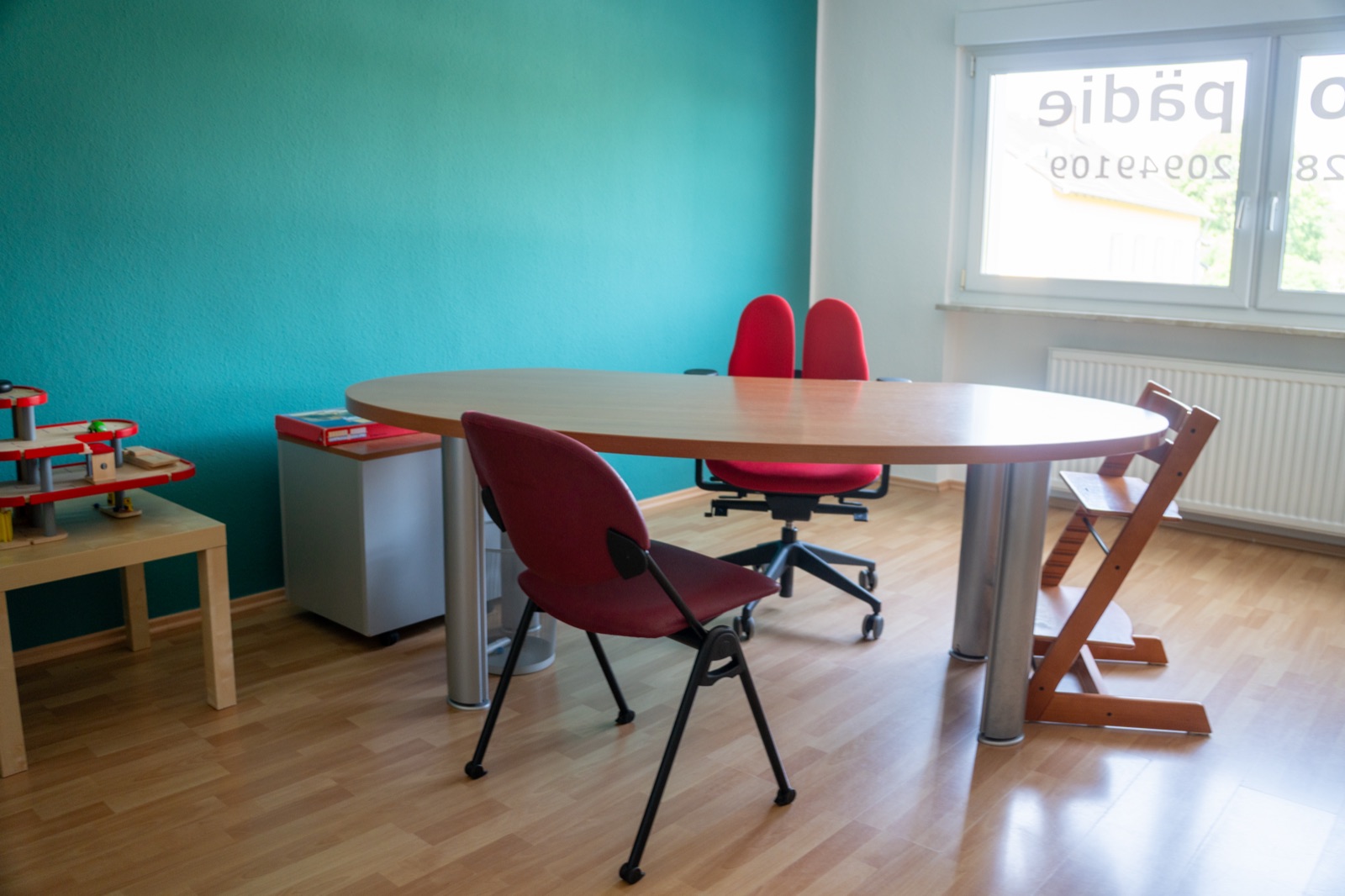Therapieraum mit ovalem Besprechungstisch, roten Stühlen und Kinderhochstuhl